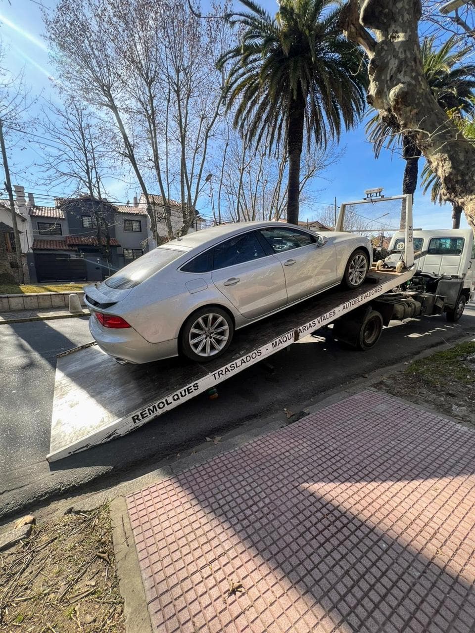 Audi A7 gris siendo cargado sobre la rampa de un camión plataforma en una calle con árboles
